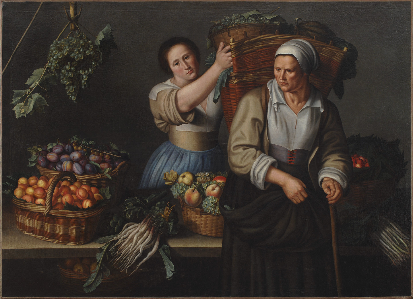 Two women are surrounded by baskets of grapes and other produce. They are both light-skinned and wearing old-fashioned attire with head wraps and full skirts, stand on either side of a table, as the younger one places a bin of grapes into a large basket on the other’s back.