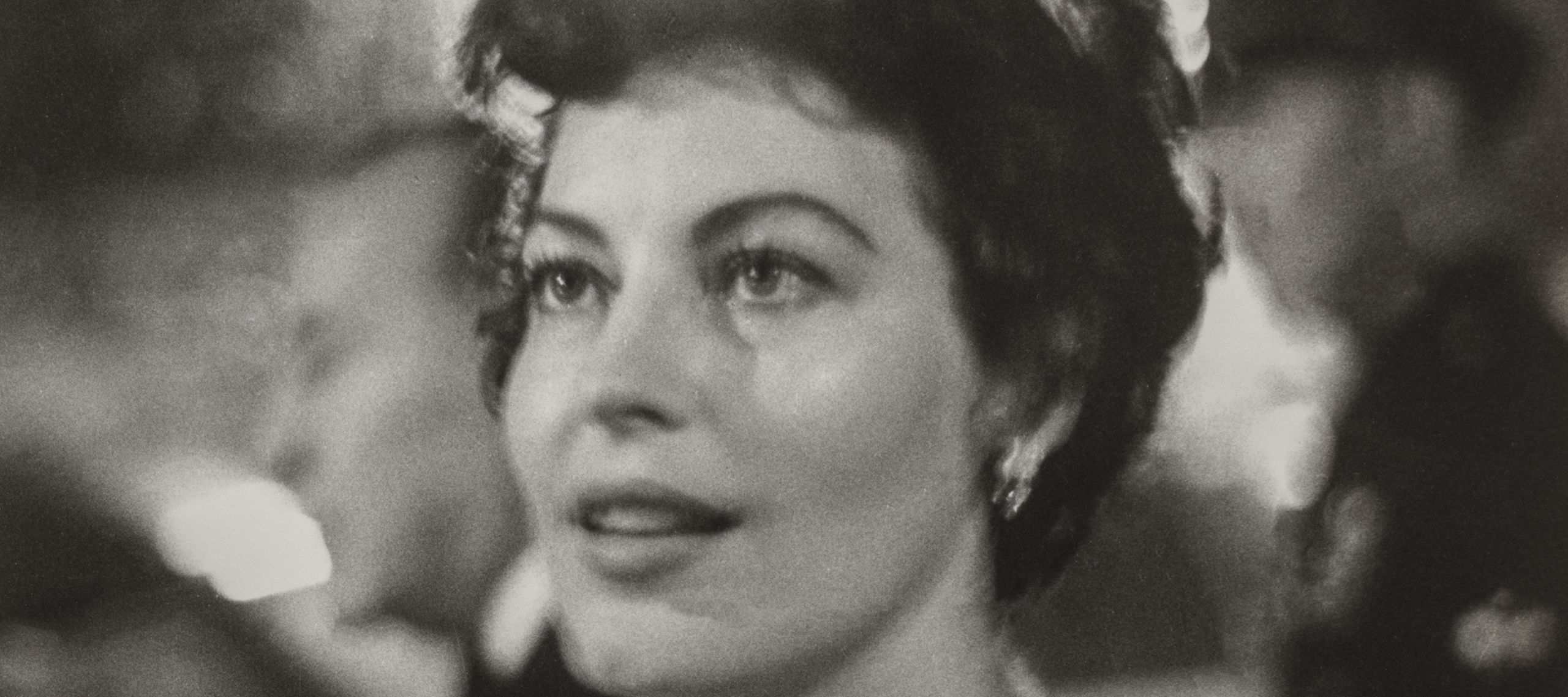 Ava Gardner, a woman with light skin tone and wavy, short brown hair, is shown from the shoulders up. She wears a jeweled necklace and is surrounded by lights and people at a party. She looks to the side of the camera at something out of frame with a fixed gaze and slight smile.