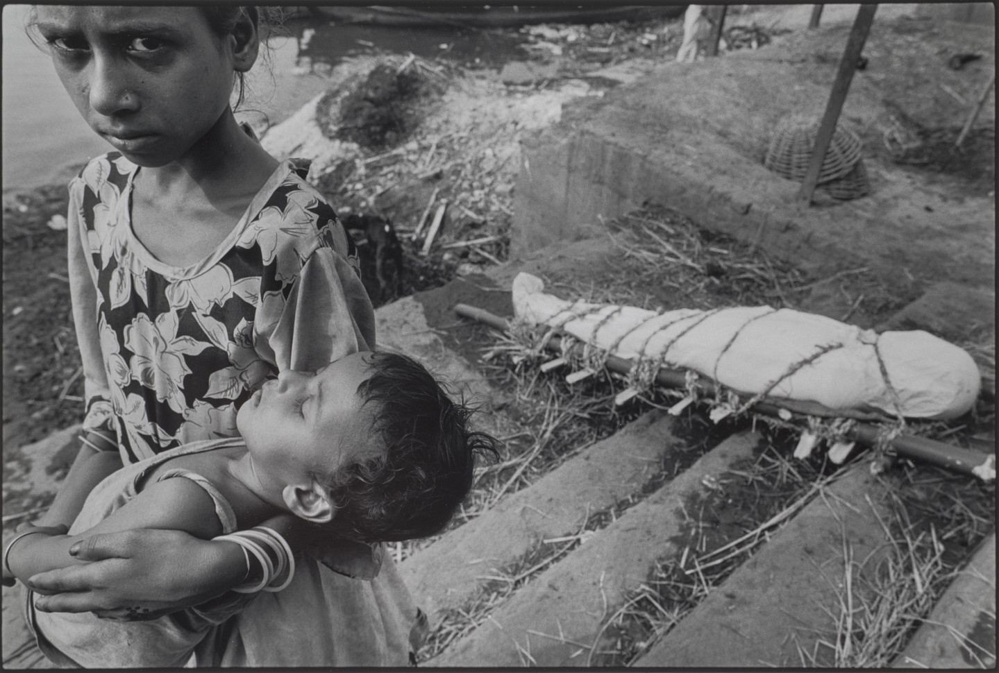 A black-and-white photograph of a child with medium skin tone carries a sleeping baby and stares into the camera with a distraught look on her face. In the background, a dead body is wrapped in white cloth and tied to a makeshift stretcher.