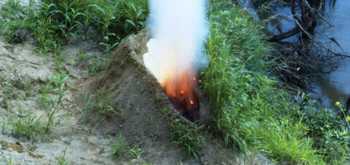 A mound of dirt in a grassy area by water. In the center of the mound is a human-shaped recess full of gunpowder that has been set aflame, exploding up in fire and white smoke, making the dirt mound resemble an erupting volcano.
