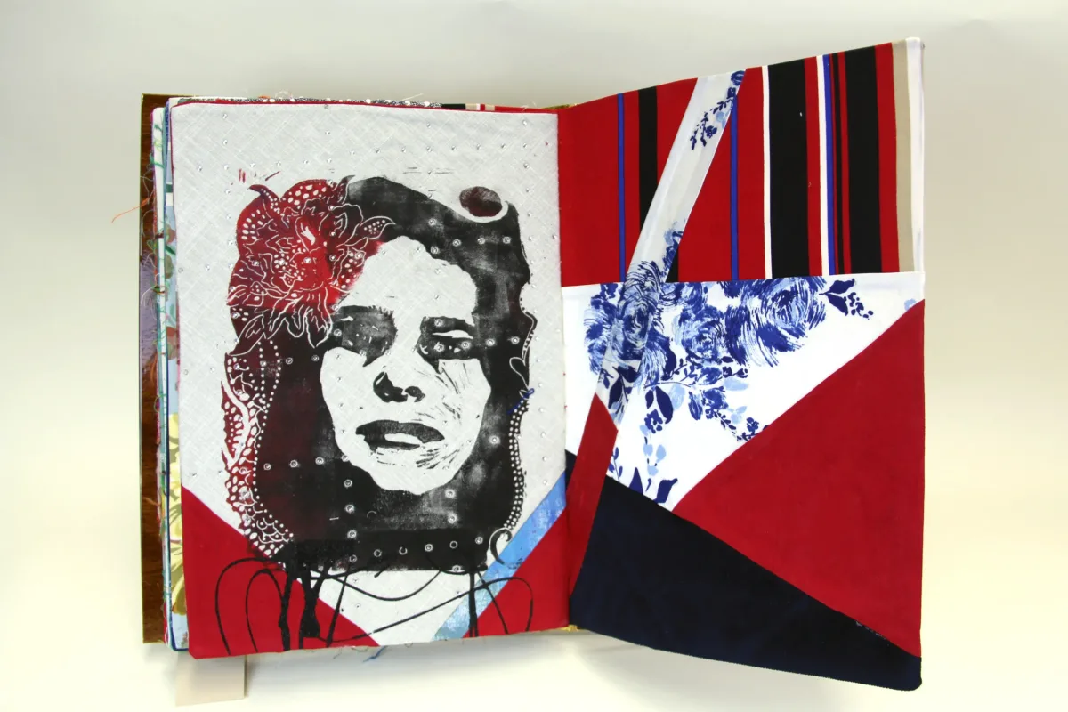 A book made from colorful print fabrics collaged and stitched together is laid flat and open to a spread that features an stencil cut image of a woman's face overlaid with an intricate flower positioned on her hair.