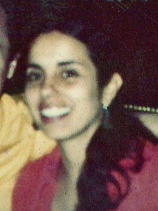 A blurry, cropped color print photograph of a medium-skinned woman with long dark hair pulled into a ponytail. She looks at the camera and smiles.