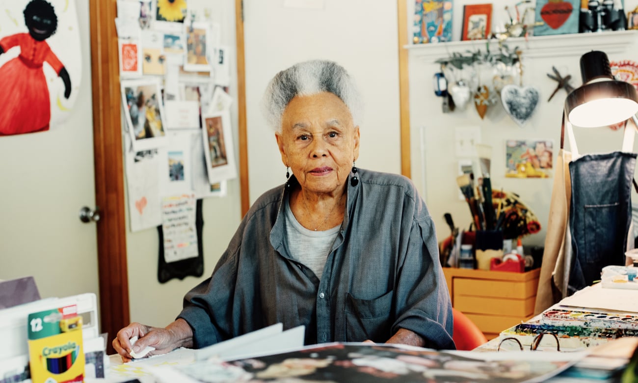 An older light-skinned African American woman sits at a table in an art studio, filled with papers hanging on the wall, craft materials, and cards and personal treasures. She stares at the camera, unsmiling but pleasant, and her white and grey hair is styled in a neat, cropped afro