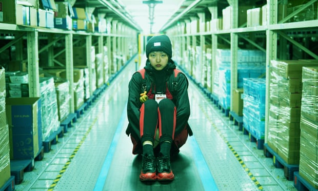 A light-skinned woman of Asian descent wears a black beanie, black jacket, and black leggings and shoes striped with red. She sits in the middle of a long, artificially-lit corridor in a warehouse, her knees bought to her chest and her hands clasped beneath them. She looks at the camera unsmiling, with an uneasy but defiant expression on her face. On either side of her, plastic-wrapped boxes on shipping palates are lined and stacked atop industrial shelving.