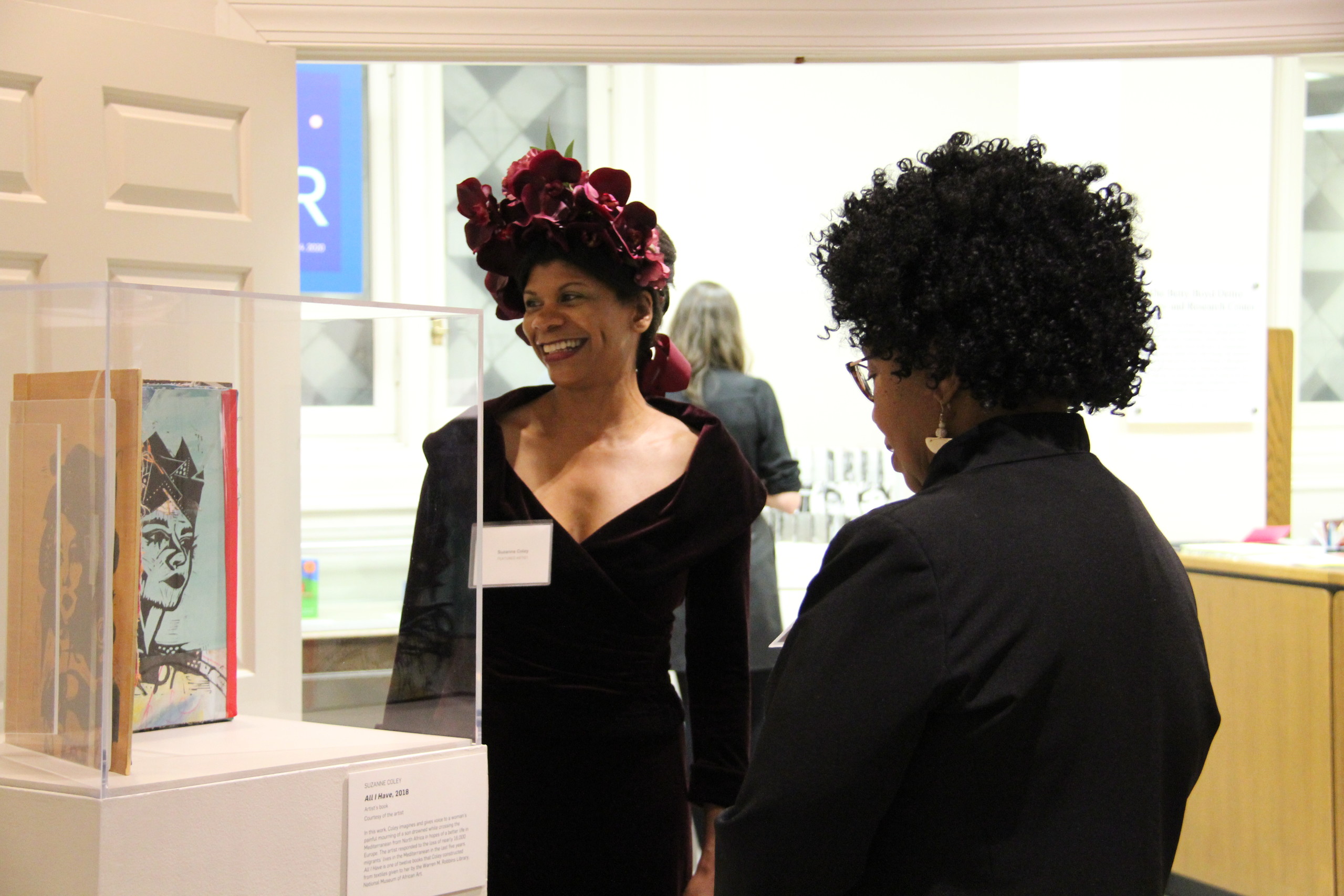 A dark-skinned woman wearing a maroon velvet floral headpiece and a velvet dress of the same color stands in front of an exhibition case smiling and looking toward the artwork in the case. Another dark-skinned woman stands with her back to the camera viewing the artwork.