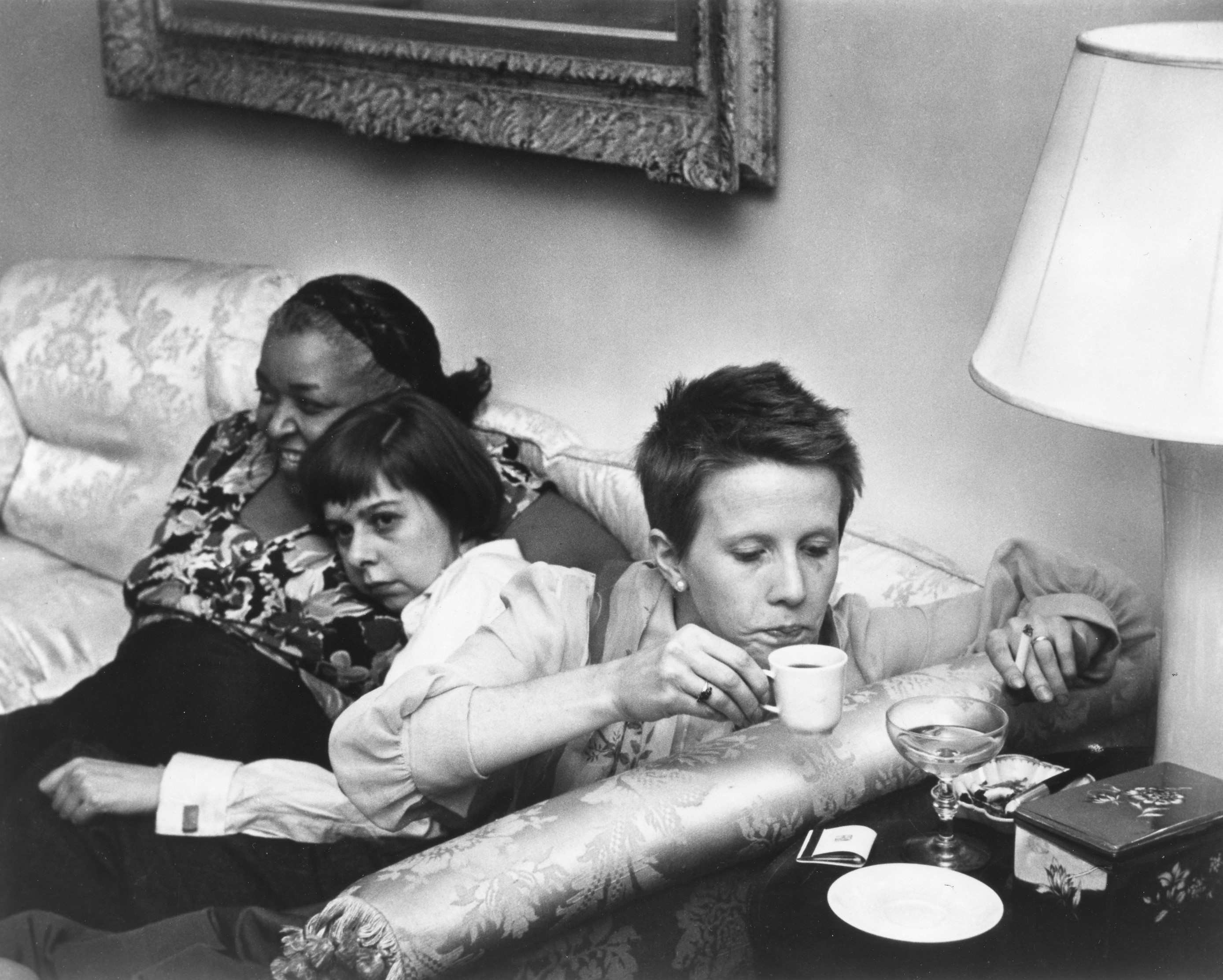 Three women sit on a couch, facing in different directions. At left, a dark-skinned woman faces out, to the left of the camera, and smiles; a light-skinned woman with bobbed dark hair leans on the first woman and gazes down. At right, a light-skinned woman with short hair leans over to lift a cup of tea and cigarette from a side table.