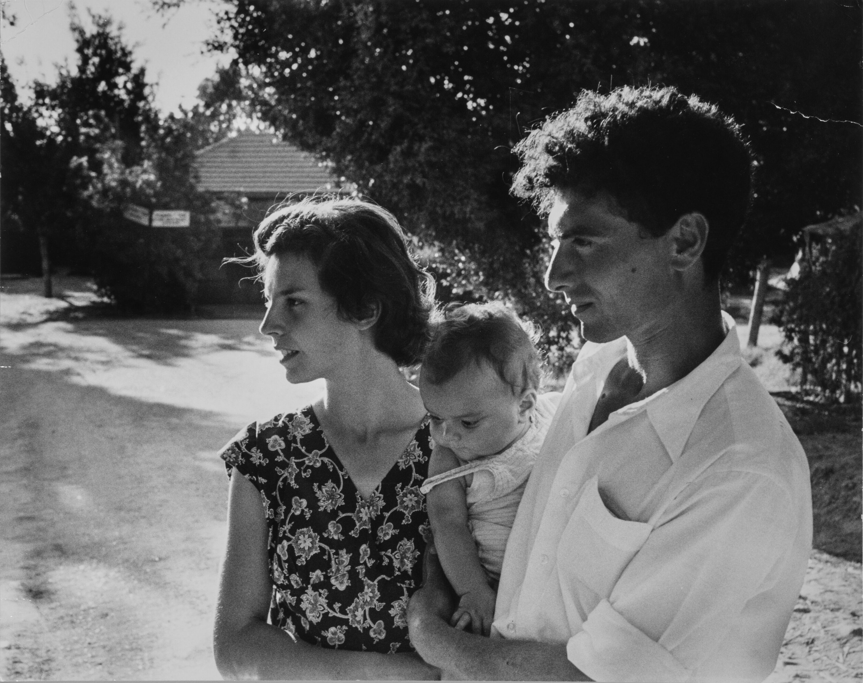 A man stands next to a woman holding a baby on his hip between the two of them; all the figures have light skin tone and brown hair. The adults are seen in profile looking to the left at something out of the frame. They are in a sunny outdoor setting in front of trees and a structure in the distance.