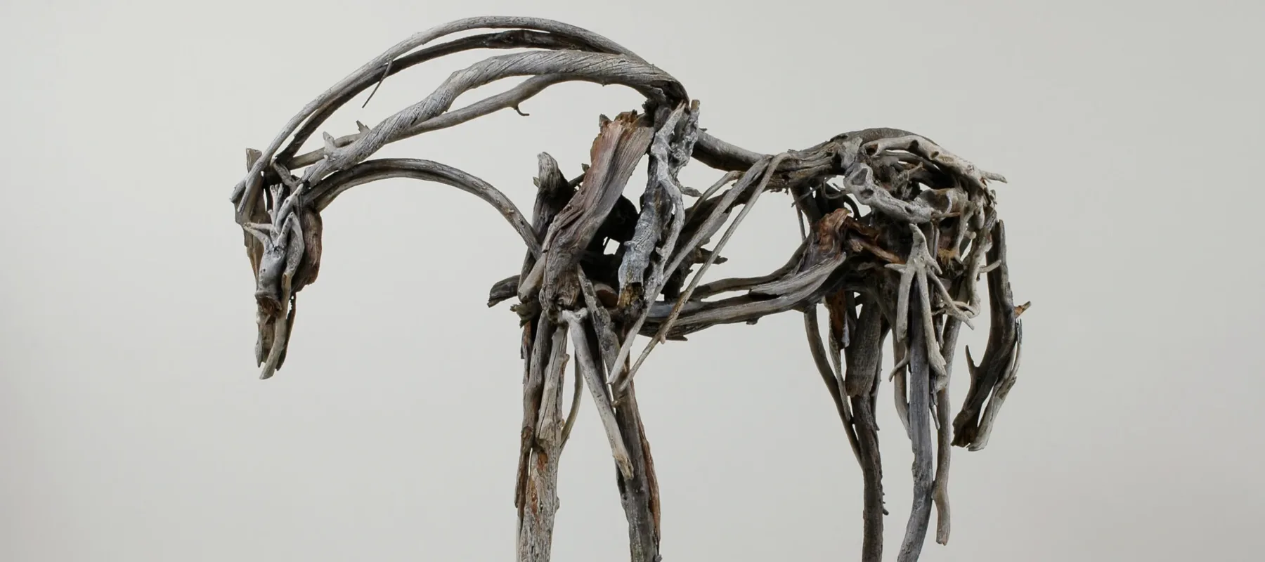 In this life-size sculpture, a horse stands facing left with its neck and head bent down. The horse appears to be made of many sticks of driftwood that are different shades of gray, different textures, and different lengths, all coming together to form a sculpture that evokes a horse’s movement.