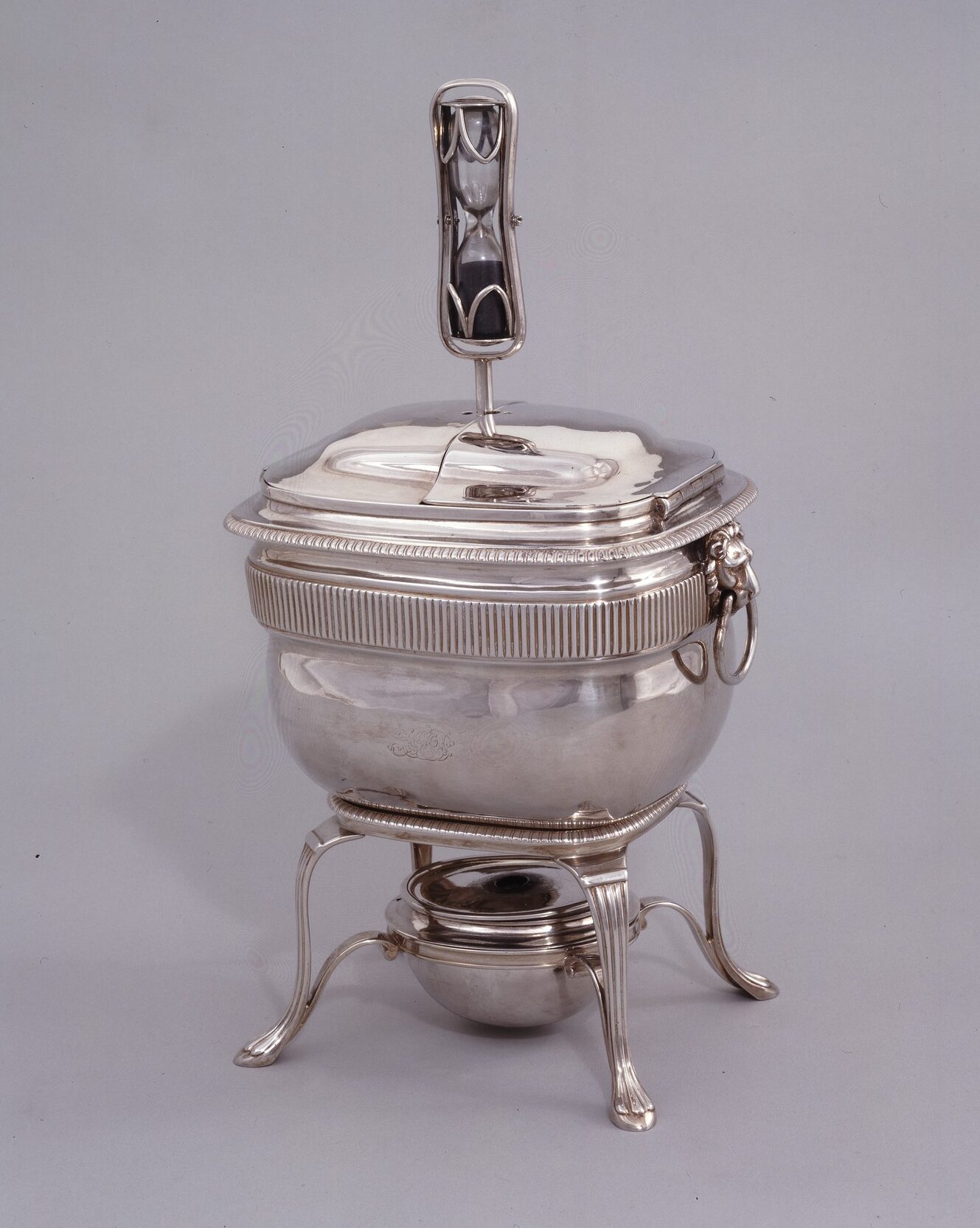 Square silver egg coddler sits on four thin legs connected to a circular burner below. A small hour-glass with black sand protrudes from the top of top.