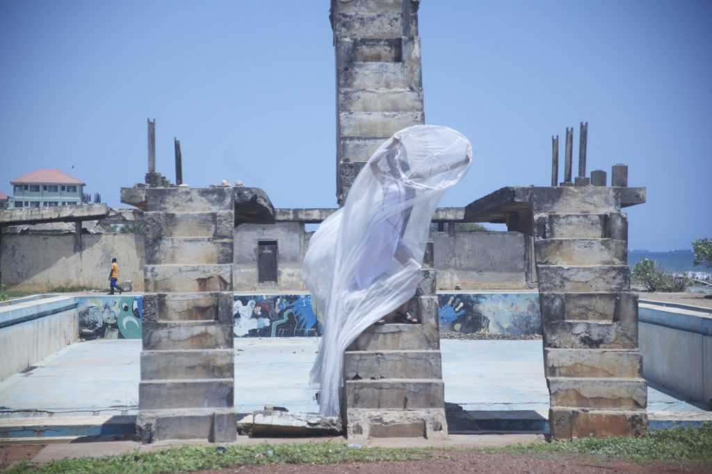A dark-skinned woman stands inside of a large white transparent fabric, posed in a dramatic fashion and wearing a white dress that covers the left side of her body. She is on the steps of an unfinished or demolished concrete building, with just it's bare elements left.