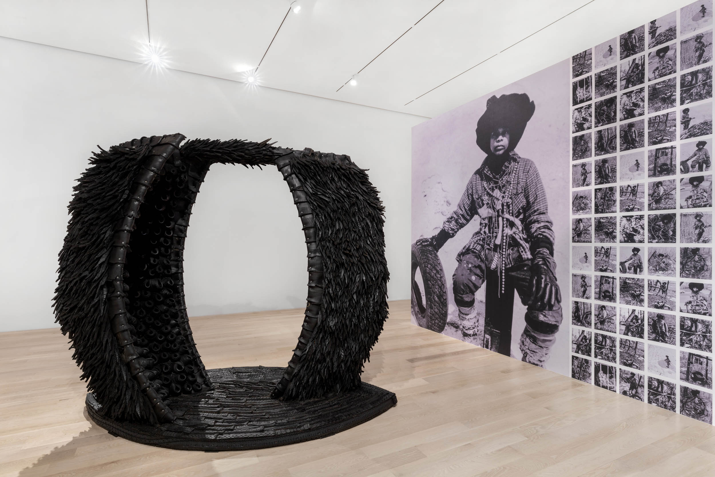 In a modern art gallery with light wood floors, a large circular sculpture of feathered rubber material sits in the middle of the room. On an adjacent wall, one large black-and-white photograph shows a dark-skinned woman in industrial dress sitting on a stool, her hand resting on a tire. Next to this photograph is an entire wall of many smaller photos featuring the same woman.