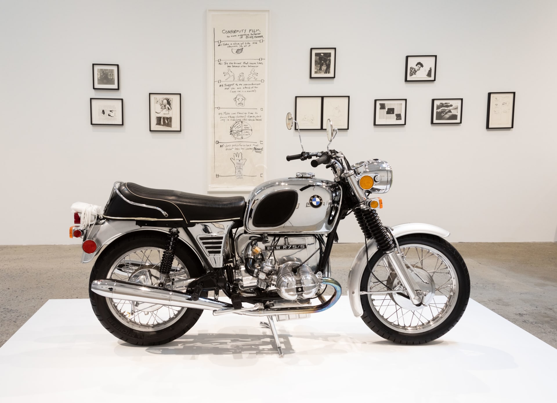 A vintage BMW motorcycle in impeccable condition sits on a white platform in the middle of a contemporary art gallery. Behind it, hanging on the wall, are a number of framed drawings, images, and journal pages.