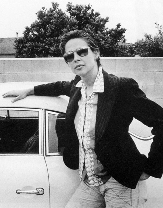 In a black-and-white photo from the 1970s, a light-skinned woman stands next to a car, her right hand resting on the hood and her left on her hip. She wears her short, brown hair slicked back, aviator sunglasses, a black blazer, and jeans. She stares confidently at the camera.