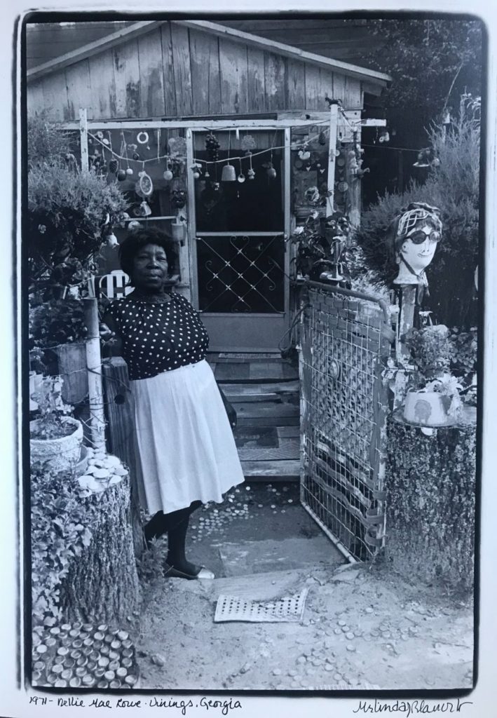 A black-and-white photo features a dark-skinned woman wearing a light skirt and polka-dotted blouse standing at the gate entrance to a small wooden house. She leans on a wooden pillar and stares confidently at the camera.