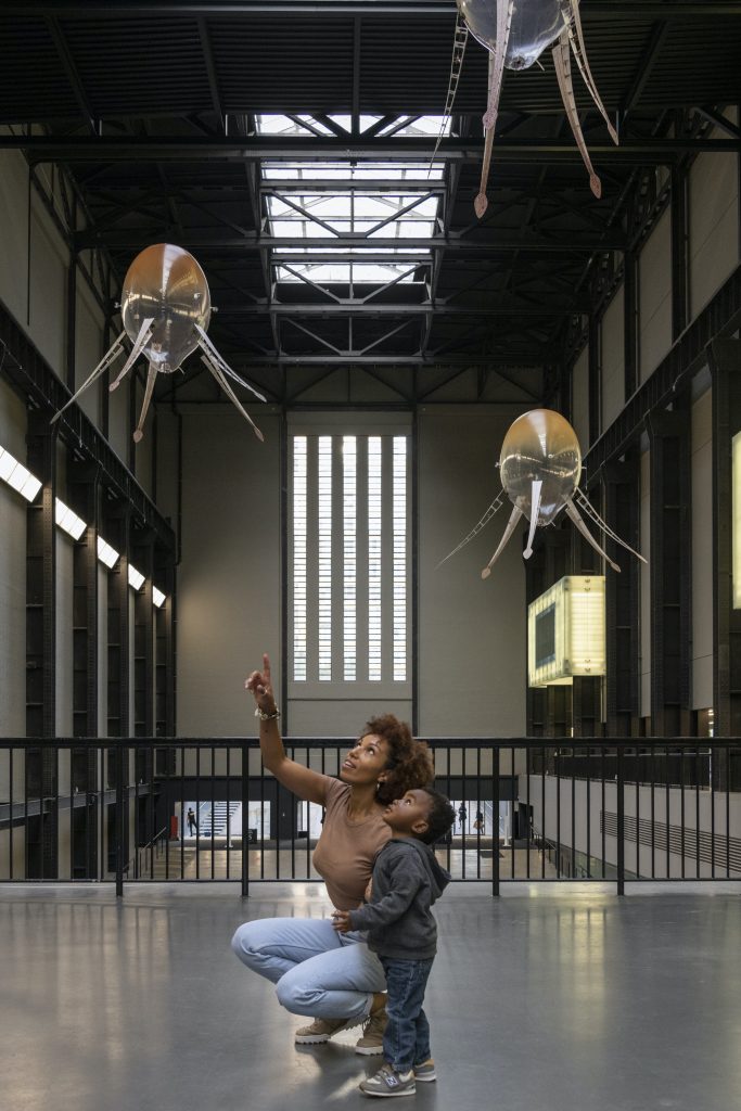 In a large industrial hall, three mechanical orbs float in the air, each with a series of "legs" splayed out. Below them, a dark-skinned woman kneels next to a toddler and points up at the orbs in wonder.
