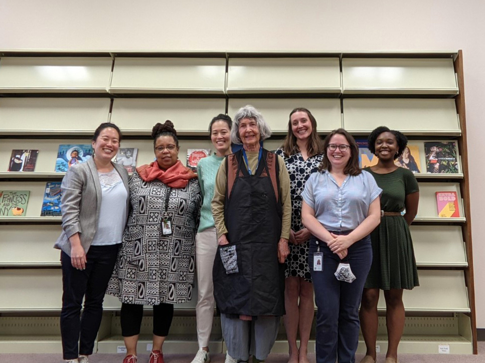 A group of seven women of various ethnic descents--Asian, African, and white--and various ages--young to older--stand smiling together in front of a tower of book display cases, which are mostly empty except for the two central racks.