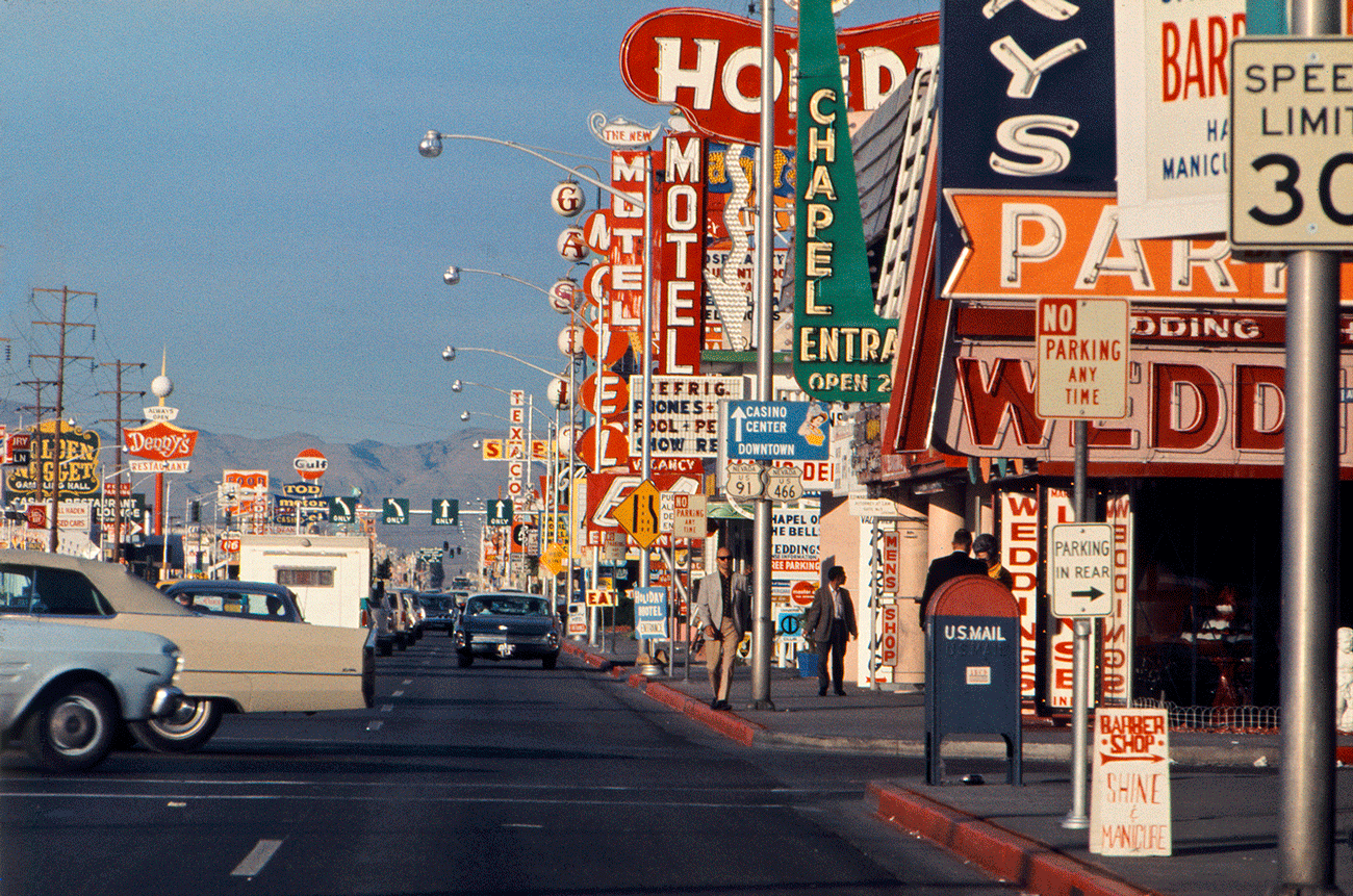 A color photograph from the 1960s shows the busy, colorful Las Vegas strip. The street is dotted with large sedans, and on the right, a strip of buildings is covered in large, multiple, signs advertising motels, wedding chapels, casinos, and more.