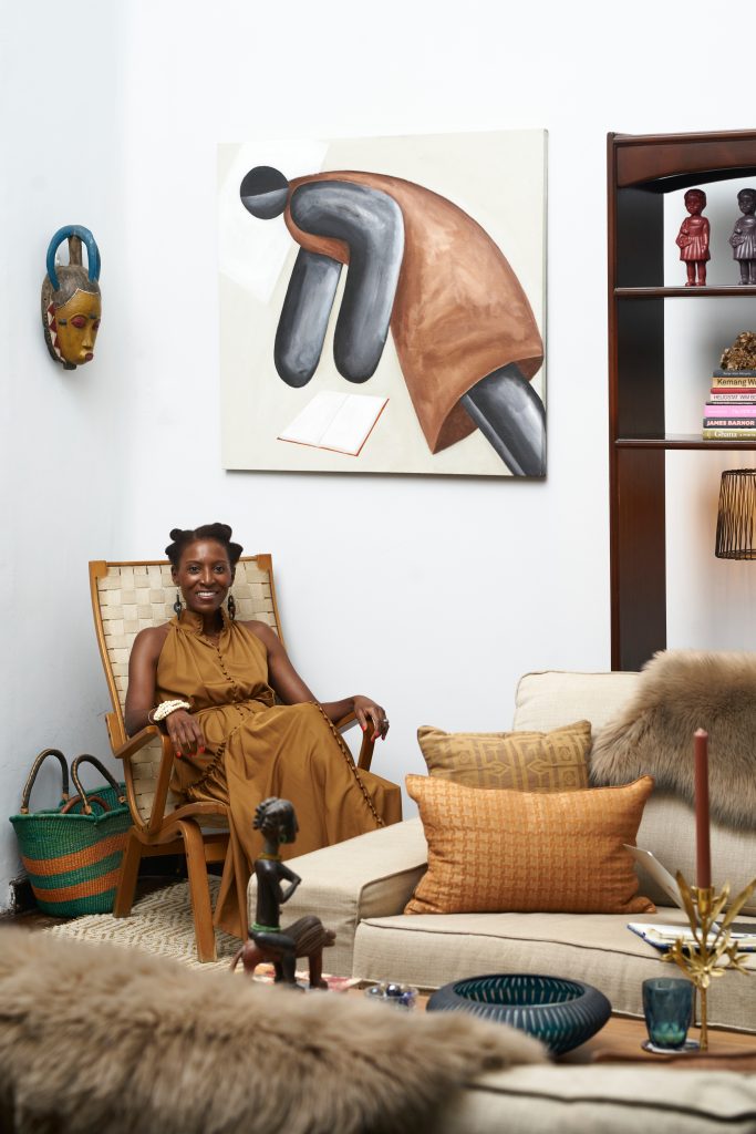 A dark-skinned woman dressed in a light brown dress with intricate buttons sits in a chair in a well-styled room. On the walls and shelves around her are different African art pieces. She smiles brightly and looks directly at the camera.