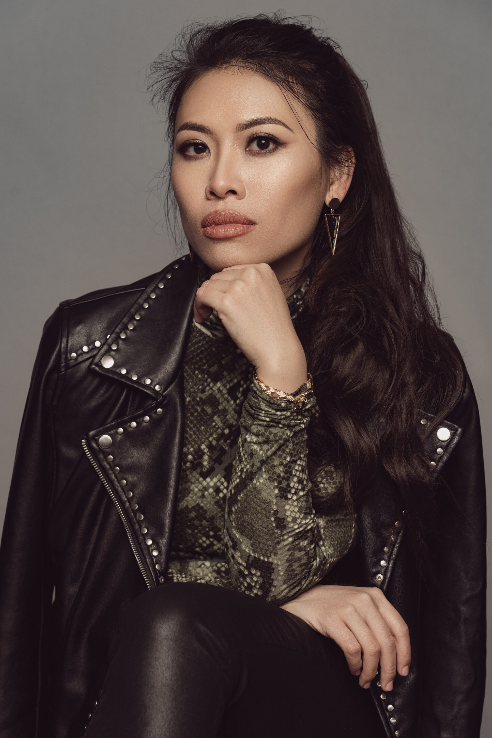 A light skinned Asian woman with long dark hair looking directly at the viewer. She is seated with her elbow resting on her knee and her chin resting on her hand. She is wearing a black leather jacket, black pants, and a green snake print shirt.