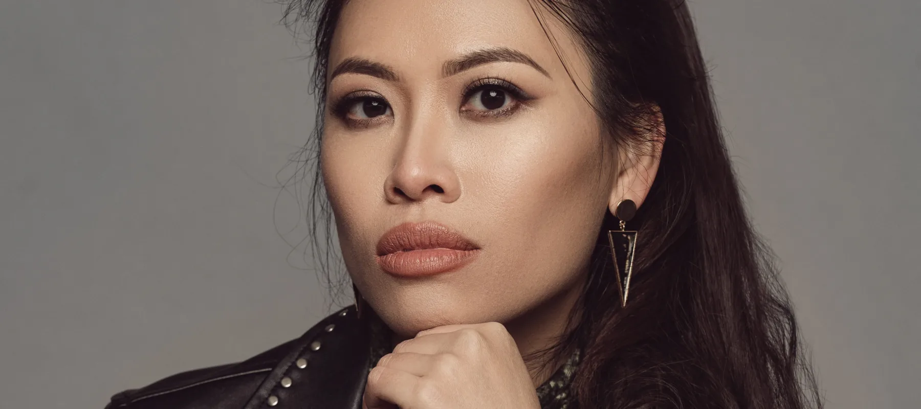 A light skinned Asian woman with long dark hair looking directly at the viewer. She is seated with her elbow resting on her knee and her chin resting on her hand. She is wearing a black leather jacket, black pants, and a green snake print shirt.