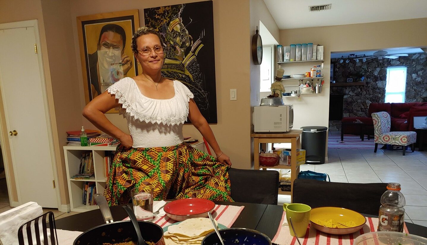 A medium skin-toned adult woman wearing traditional Puerto Rican dress—a white blouse with lace neck and brightly patterned skirt—stands smiling in front of her dining table in her home. The table is set with bowls and pots of ground beef, salsa, sauce, guacamole, and salad.
