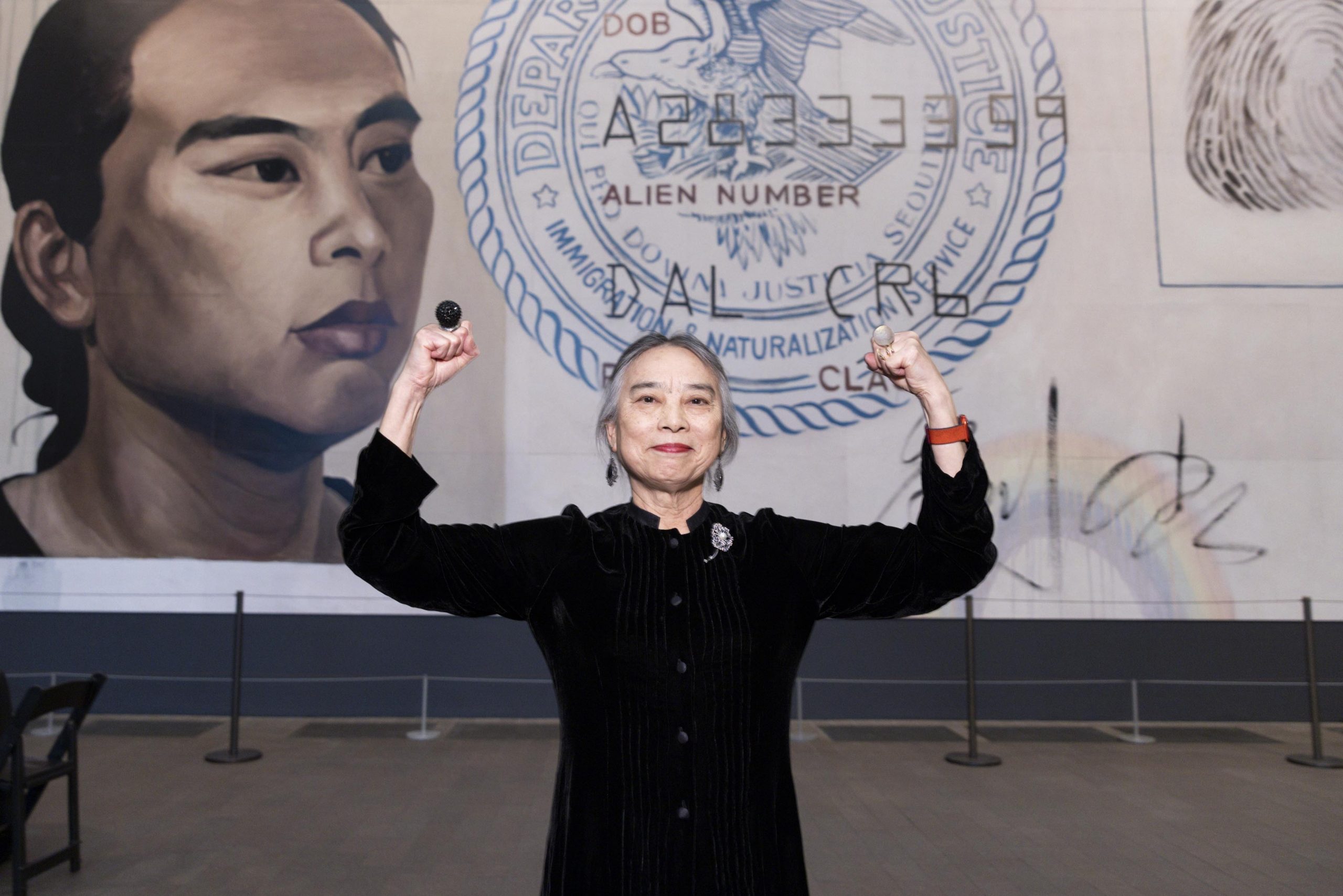 A light-skinned woman of Asian descent stands in front of a large-scale painting of a United States immigration/naturalization identification card. She wears a black button-up dress and stands with both of her arms raised and flexed, in a bicep curl. She smiles slightly. Two large, globular rings on either hand stand out.