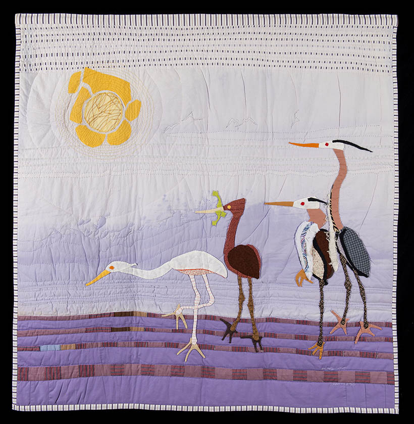 A quilt features a scene of four different colored, long-legged birds walking across the ground, as if they are looking for something. One holds something in its mouth. A bright yellow sun is sewn in the upper left corner.