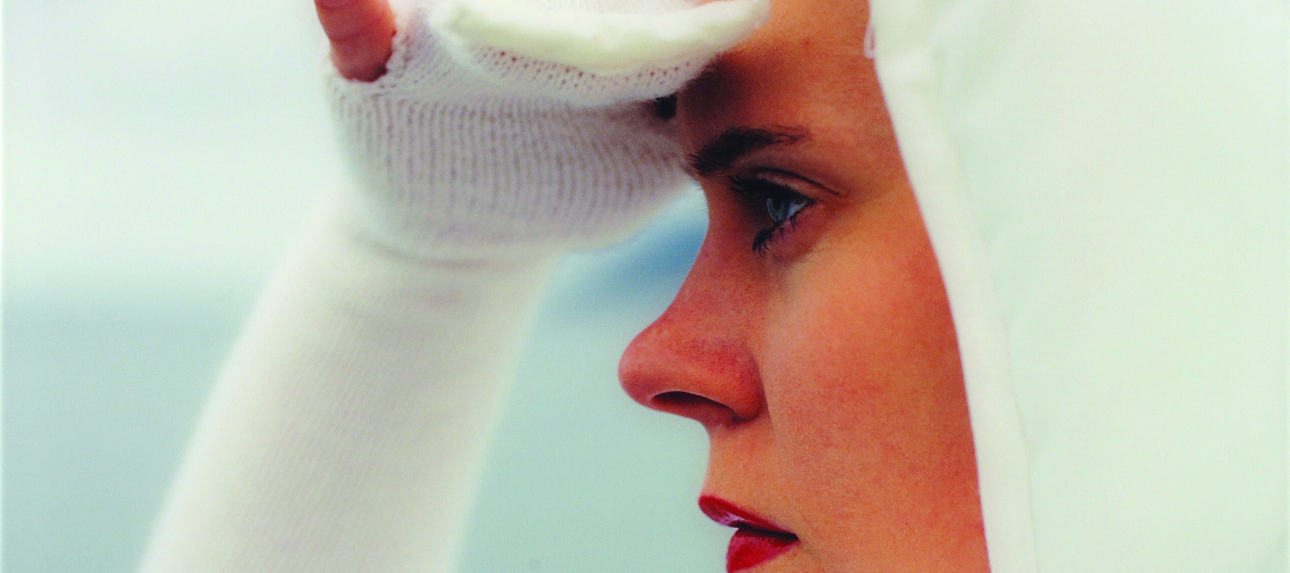 Square close-up of a light-skinned woman in profile looking to the left, seen from the shoulders up. She wears all white, including a bonnet and knit sleeves that become gloves covering her three middle fingers, and she holds her right hand up as if shielding her eyes from the sun.