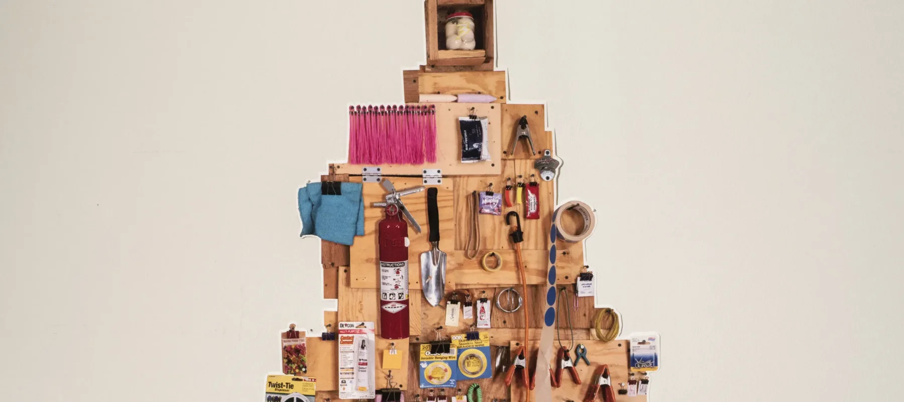 Wooden rectangles in different shades and sizes arranged in the shape of a pine tree, covered in in a colorful, eclectic, hanging assortment of common household and gardening tools—such as a trowel, rolls of tape, gardening gloves, a fire extinguisher, rulers, and more.