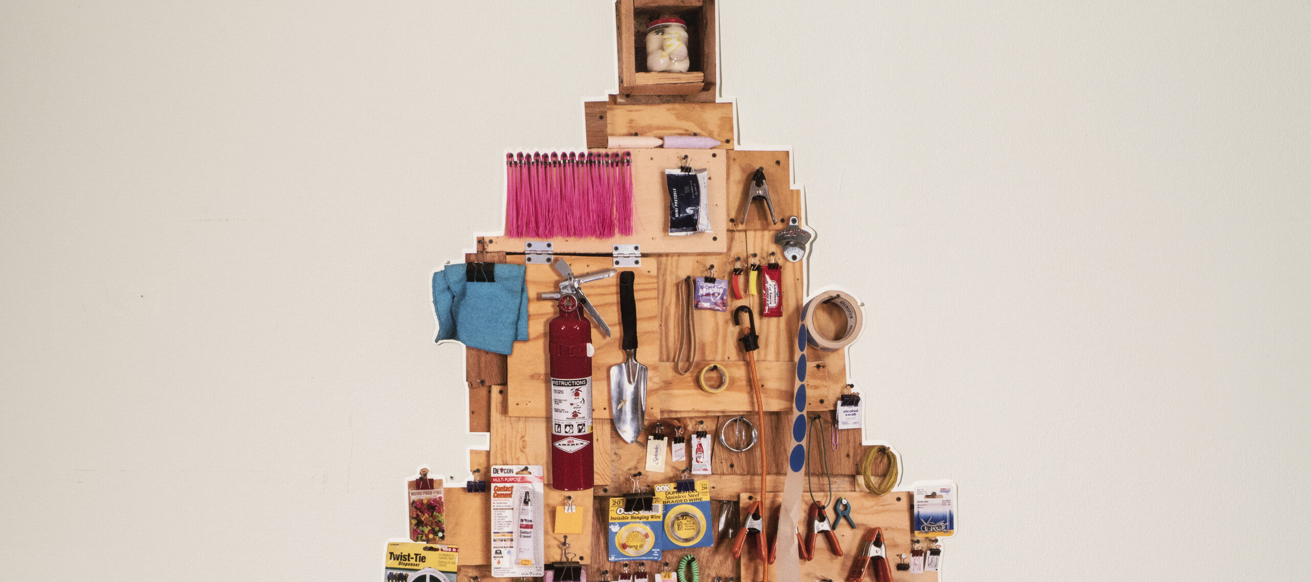 Wooden rectangles in different shades and sizes arranged in the shape of a pine tree, covered in in a colorful, eclectic, hanging assortment of common household and gardening tools—such as a trowel, rolls of tape, gardening gloves, a fire extinguisher, rulers, and more.