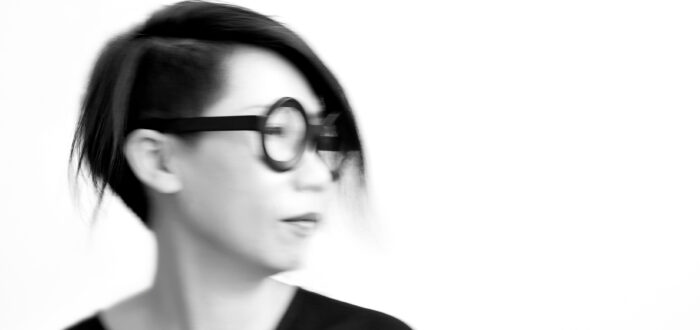 A black-and-white photo shows the blurry portrait of a light-skinned woman of Asian descent looking off to the right. She wears her dark hair cropped close and short, wide-rimmed circular glasses, and a black V-neck shirt.