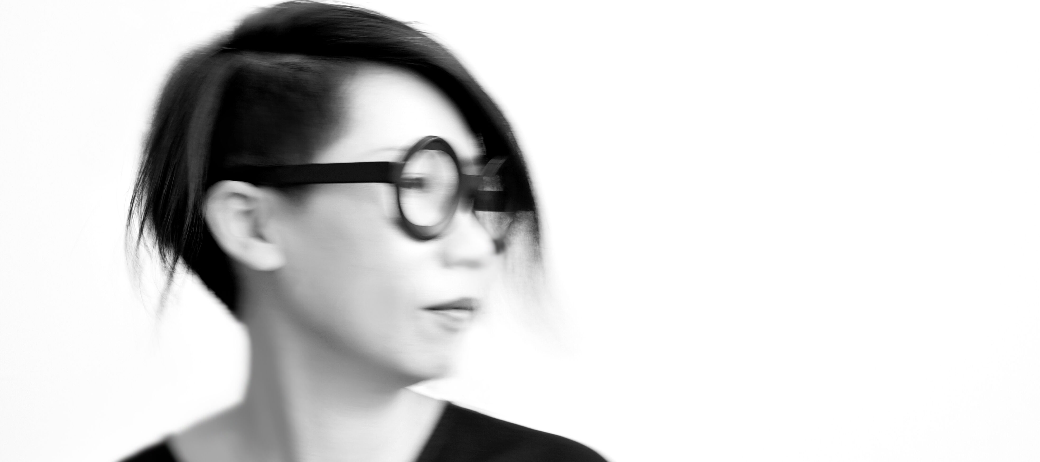 A black-and-white photo shows the blurry portrait of a light-skinned woman of Asian descent looking off to the right. She wears her dark hair cropped close and short, wide-rimmed circular glasses, and a black V-neck shirt.