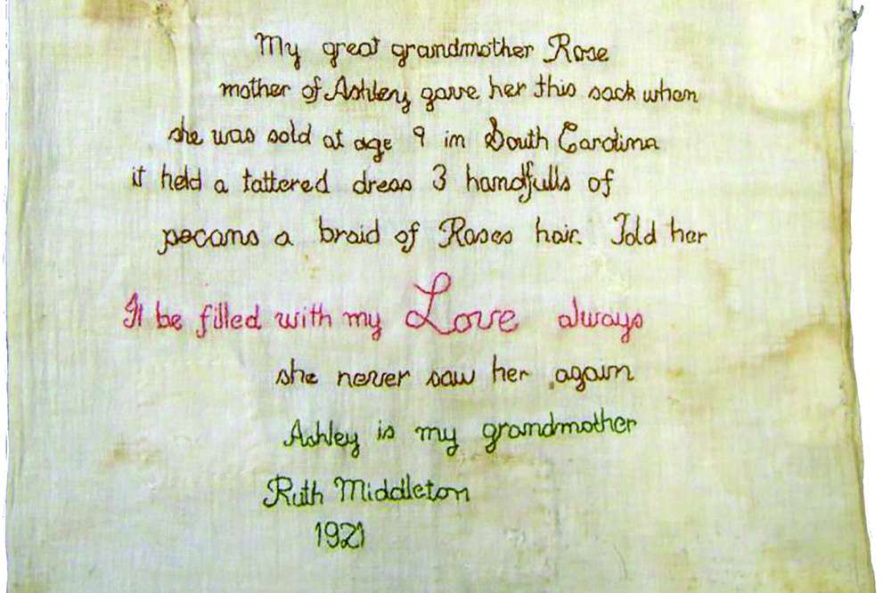 An aged, white cotton sack is embroidered with the history of the item in brown, pink, and green cursive. It reads: "My great grandmother Rose, mother of Ashley, gave her this sack when she was sold at age 9 in South Carolina. It held a tattered dress, 3 handfulls of pecans, a braid of Rose's hair. Told her "It be filled with my Love always." She never saw her again. Ashley is my grandmother. Ruth Middleton. 1921.