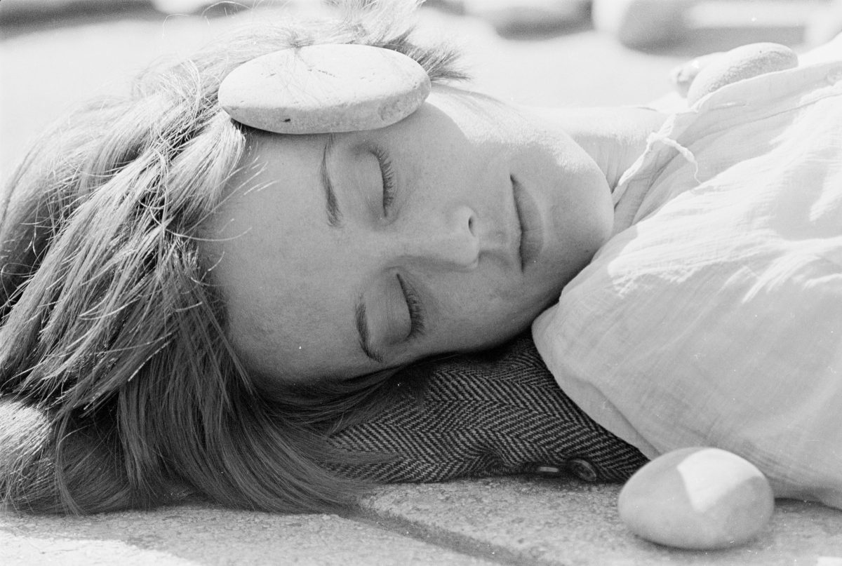 A close-up, black-and-white photo of a light-skinned woman's face and neck. She is laying on a pillow, with her head turned towards the camera, her eyes closed, and a medium-sized flat stone resting on her temple. Another stone is placed by her shoulder. She looks peaceful.