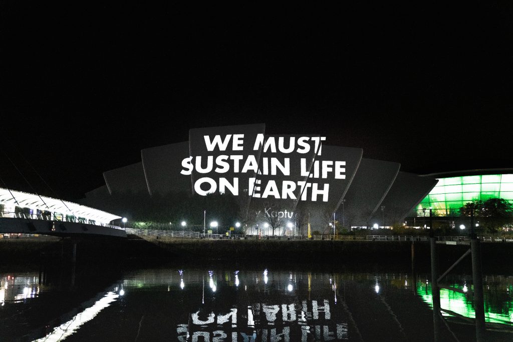 In capitalized all-white letters, the message "WE MUST SUSTAIN LIFE ON EARTH" is projected onto the exterior of the Sydney Opera House at night.
