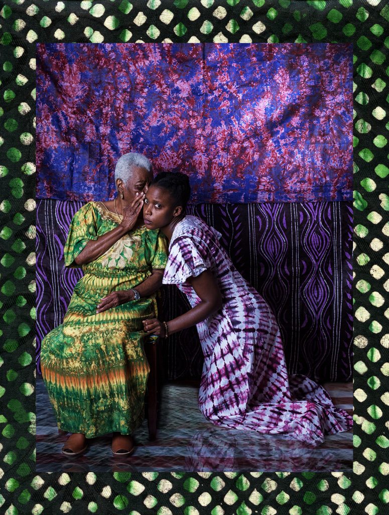 An older dark-skinned woman with white, cropped hair sits in a chair and leans towards a younger dark-skinned woman kneeling on the floor next to her, whispering something in her ear. The older woman's face is obscured by her hand, which she holds up to her mouth. The younger woman looks suspiciously towards the camera. Both wear vibrant patterned dresses. Behind them, the wall is covered in two textiles featuring pink and purple floral and patterned designs.