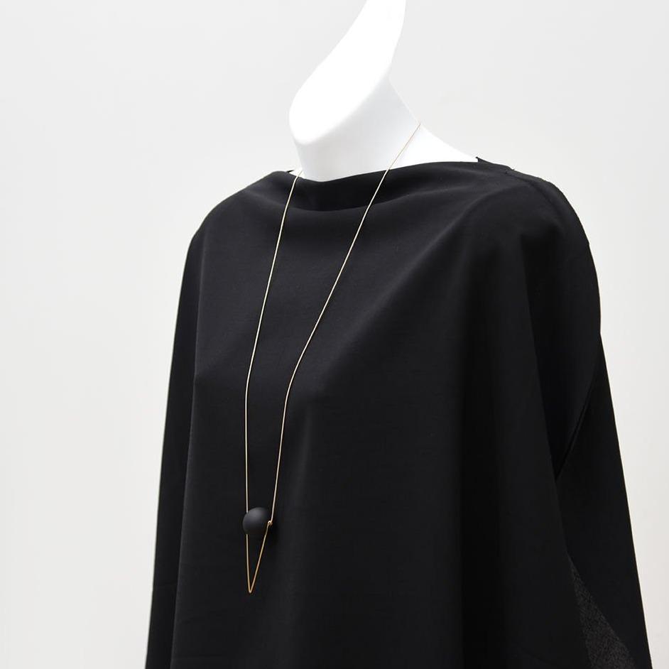 A white mannequin without a head is draped in a black shawl against a white background. A long, modern, minimalist necklace hangs around the mannequin's neck, featuring a thin brass chain that culminates in a triangle shape marked by a black matte orb.