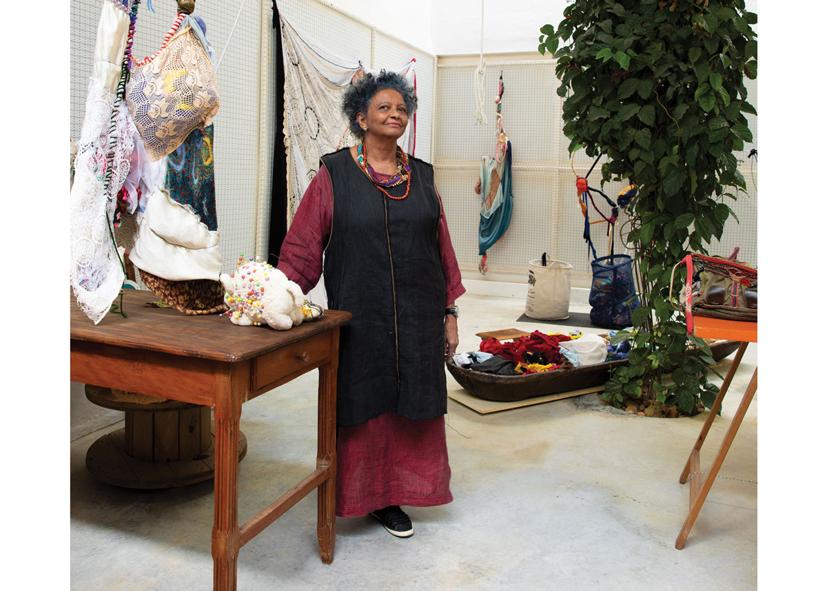A dark-skinned woman with short, curly black-grey hair stands smiling in the middle of a clean art studio with white walls and floors. In the middle of the room a tree emerges from the floor. Different sized and shaped textiles hang on the walls and are bundled on the floor.