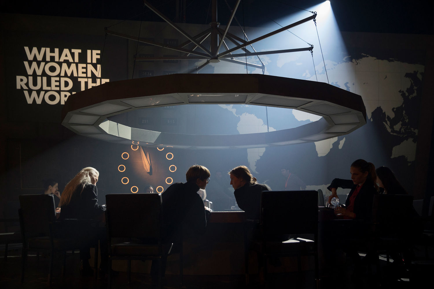 A photograph of a darkened room, with only natural, diffused light coming from the ceiling illuminating a table around which six women sit and talk and work. One pours water. They are dressed formally. A large map of the world is on the wall in front of them, along with the question, presented in bold white text, "What if Women Ruled the World?"
