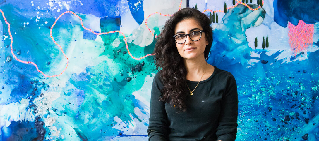 A light skinned woman with long curly dark hair wearing a long sleeved black t-shirt, grey jeans covered in paint splatters, navy blue Converse sneakers, and dark framed glasses, sits on a metal stool in front of a colorful abstract painting with large blue and green areas of paint, small green trees lining the pools of paint, and pink ropes with yellow tassels weaving across the top of the canvas.