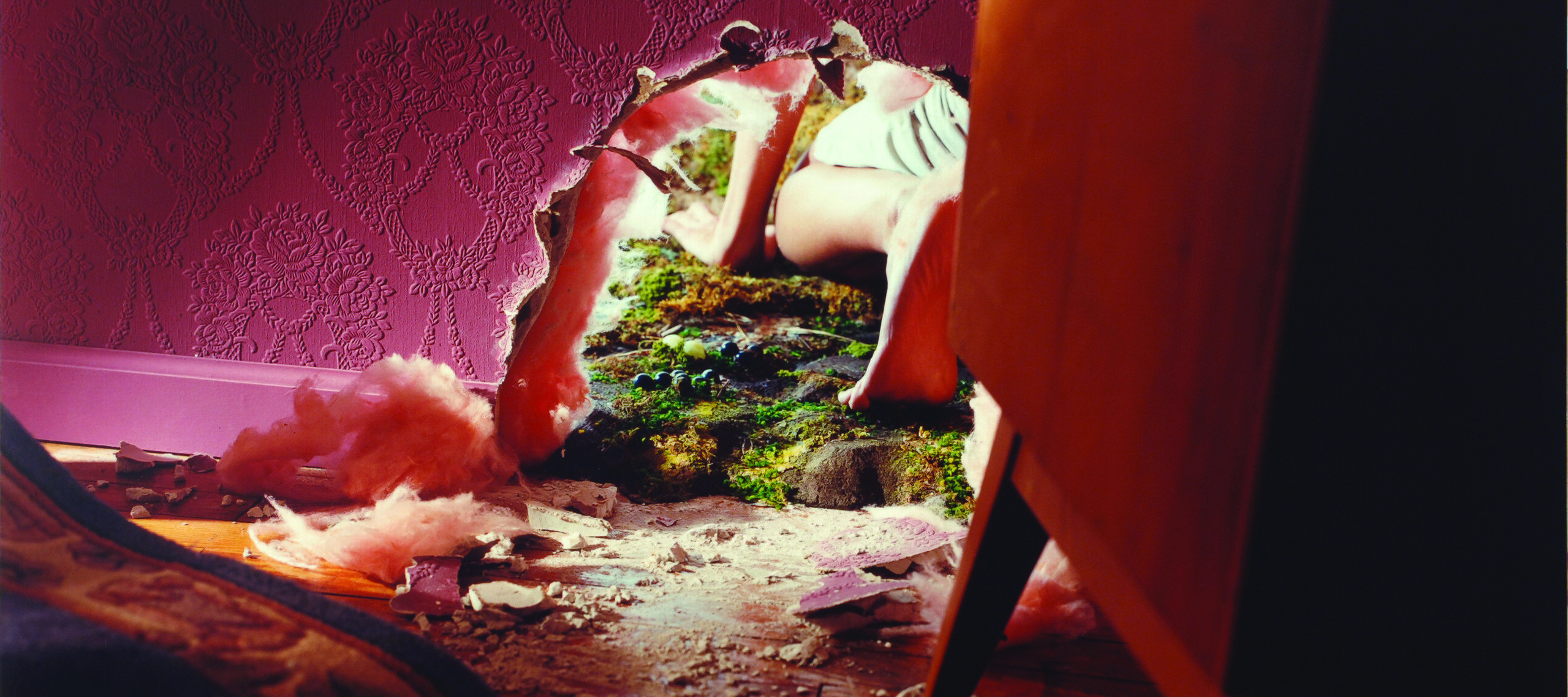A pink wall and pink chair in a living room with a hole torn through the wall at the ground-level. A child crawls out and away from the hole into the grassy outdoors, leaving behind wall debris in the room. Only the child's left arm and leg are visible through the hole.