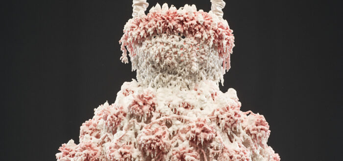 Myriad layers of melted pink and white wax encrust and obscure the metal armature for this abstract sculpture, which hangs from satin-wrapped chains. Its color and shape, as well as the bumpy, lacy texture, evoke a frilly tutu, lavishly frosted wedding cake, or coral accretions.