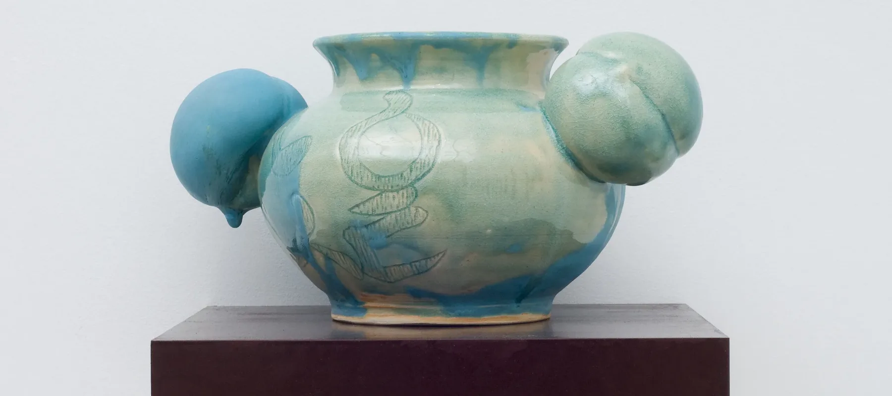 A round, ceramic vessel glazed unevenly in glossy celadon has a bulging body incised with abstract shapes. Matte turquoise glaze dribbling down from the rim obscures portions of the pale green and fully coats one of two peach-shaped orbs projecting from either side of the neck.