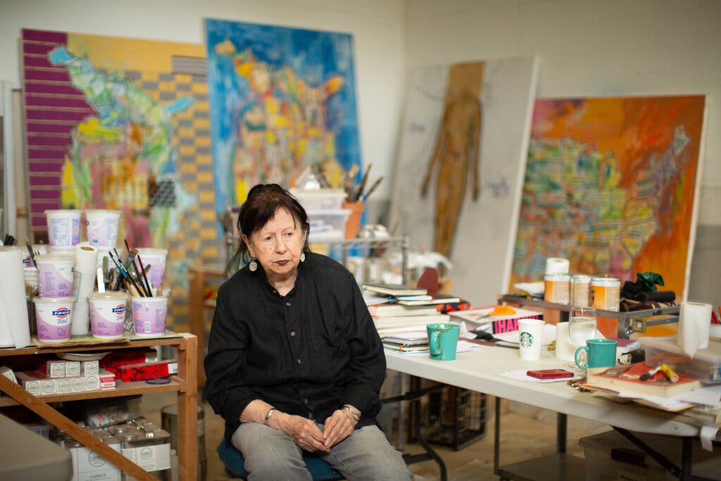 A portrait of an older, light-skinned Native American Woman sitting on a chair in an art studio. Behind her, four large canvases are propped against the wall, Tables and shelves filled with supplies and brushes. She looks pensive and stares to the right of the frame.