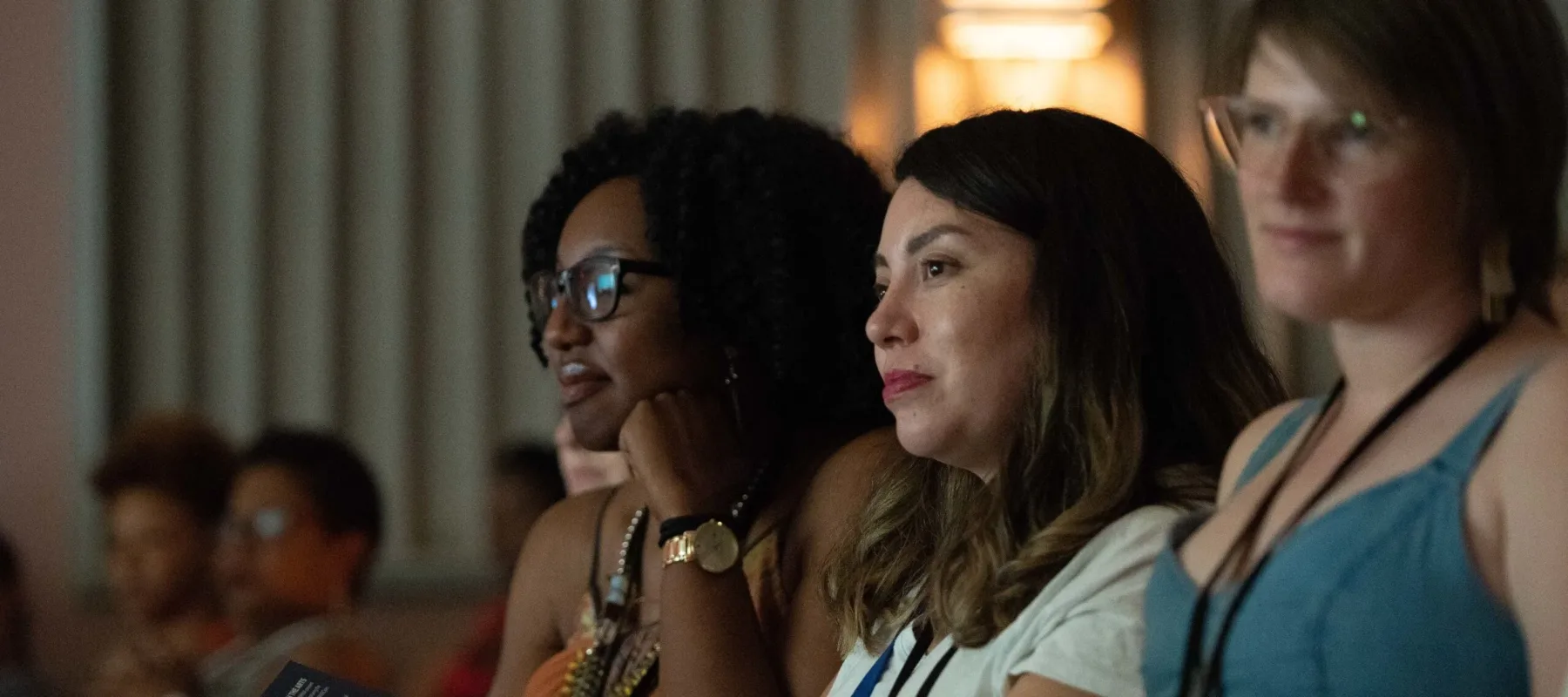 Two light skinned women and one dark skinned woman sit in an auditorium watching a performance.