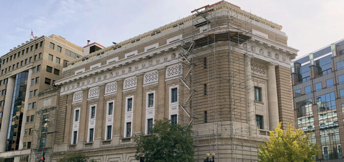 The National Museum of Women in the Arts’ historic Renaissance Revival building is pictured on a sunny day in downtown Washington, D.C. Scaffolding is set up on the front corner of the wedge-shaped structure, from top to bottom. On the sidewalk below, there is a covered walkway, panels of wood covering the doors and windows, and orange-and-white construction barriers.