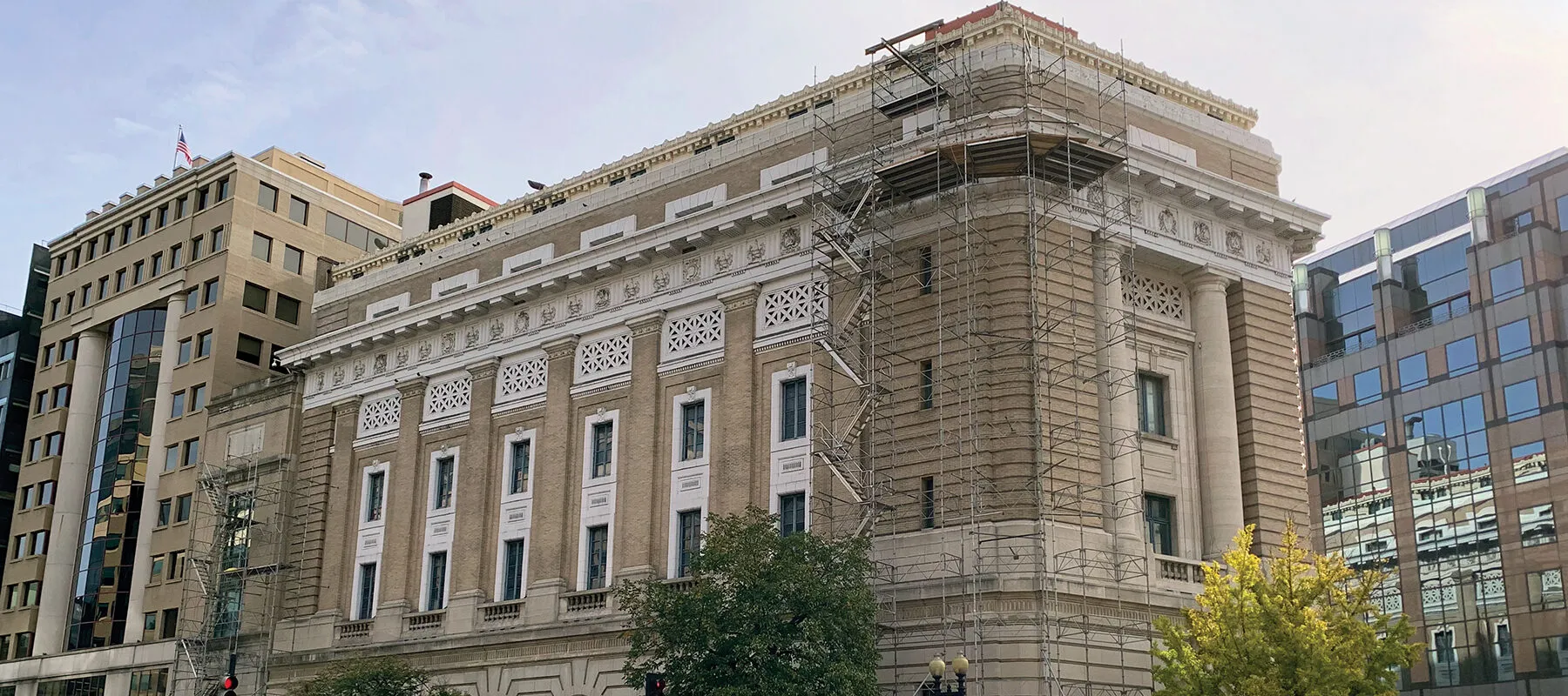 The National Museum of Women in the Arts’ historic Renaissance Revival building is pictured on a sunny day in downtown Washington, D.C. Scaffolding is set up on the front corner of the wedge-shaped structure, from top to bottom. On the sidewalk below, there is a covered walkway, panels of wood covering the doors and windows, and orange-and-white construction barriers.