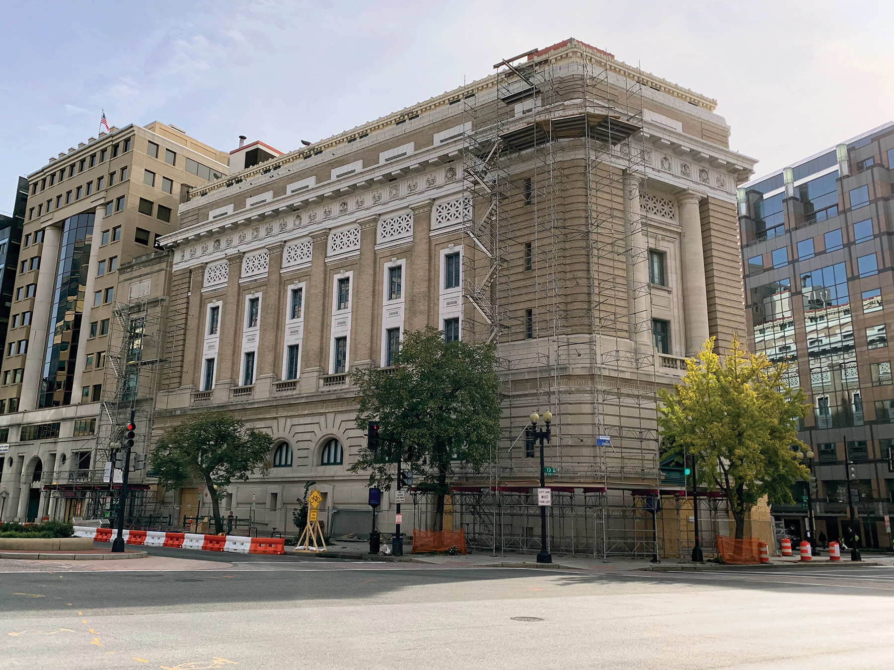 The National Museum of Women in the Arts’ historic Renaissance Revival building is pictured on a sunny day in downtown Washington, D.C. Scaffolding is set up on the front corner of the wedge-shaped structure, from top to bottom. On the sidewalk below, there is a covered walkway, panels of wood covering the doors and windows, and orange-and-white construction barriers.