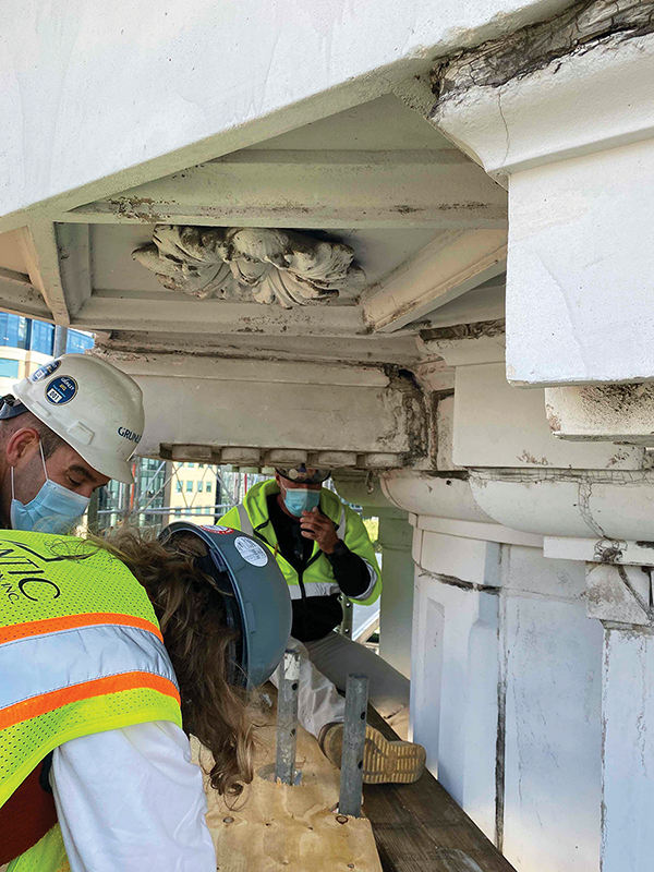 Three construction workers wearing hard hats, yellow vests, and blue medical face masks inspect the museum building’s white historic cornice. It shows signs of age, with chipped paint, weathering, and dirty areas.