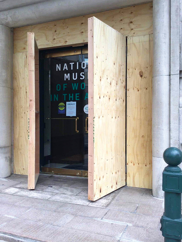 The front entrance of the National Museum of Women in the Arts is shown covered in light plywood. Two large panels, which cover the doors, are slightly ajar, revealing the building’s glass doors bearing the museum’s logo, along with several smaller signs.